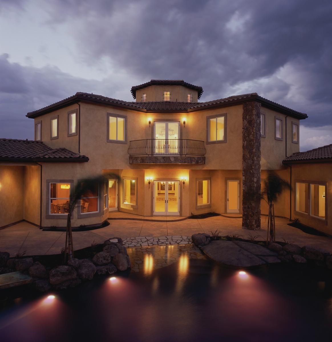 Luxury villa at dusk with warm lit windows, balcony, and reflections in a pool