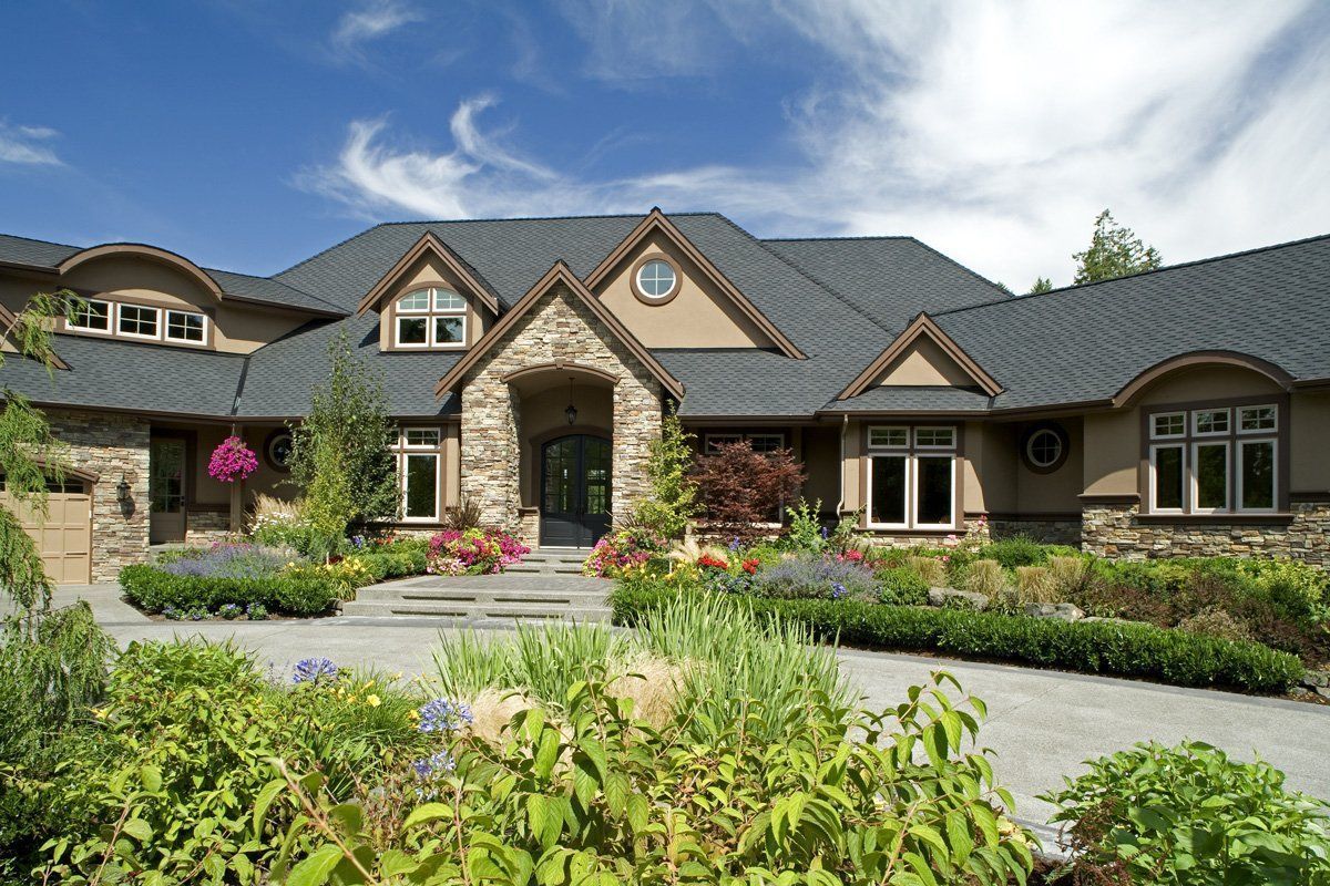 Large stone-and-wood house with landscaped garden and paved driveway under a blue sky