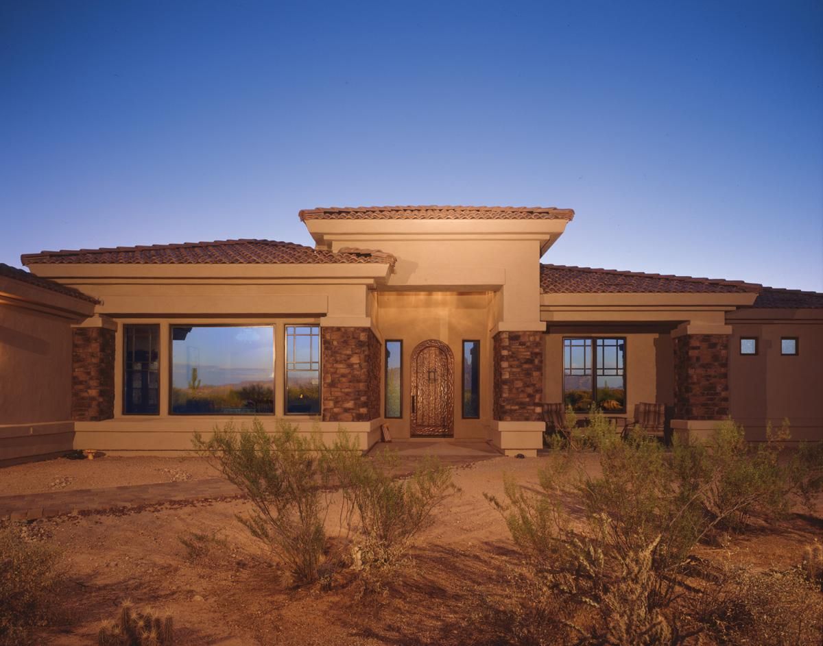 Adobe-style desert house at dusk with tan stucco, stone columns, and sparse shrubs in front
