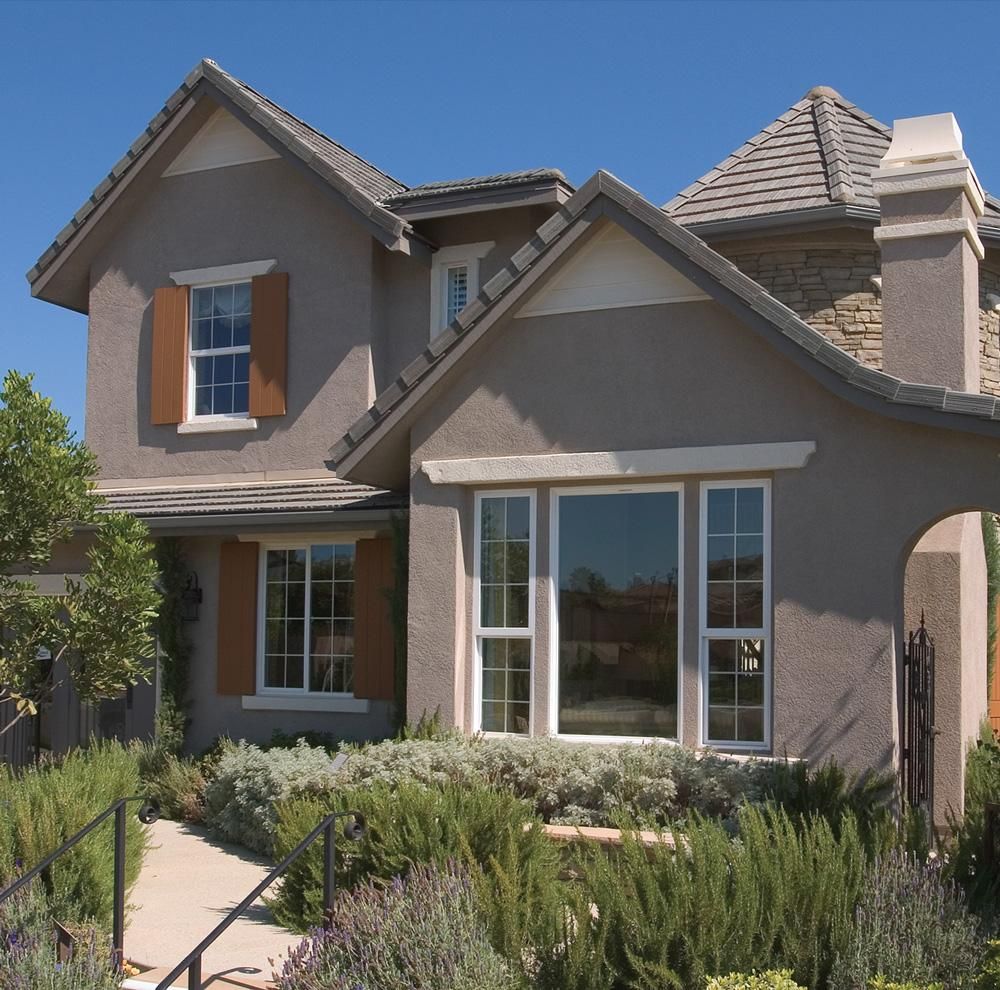 Two-story stucco house with tan trim, white windows, and a landscaped front yard under a clear blue sky