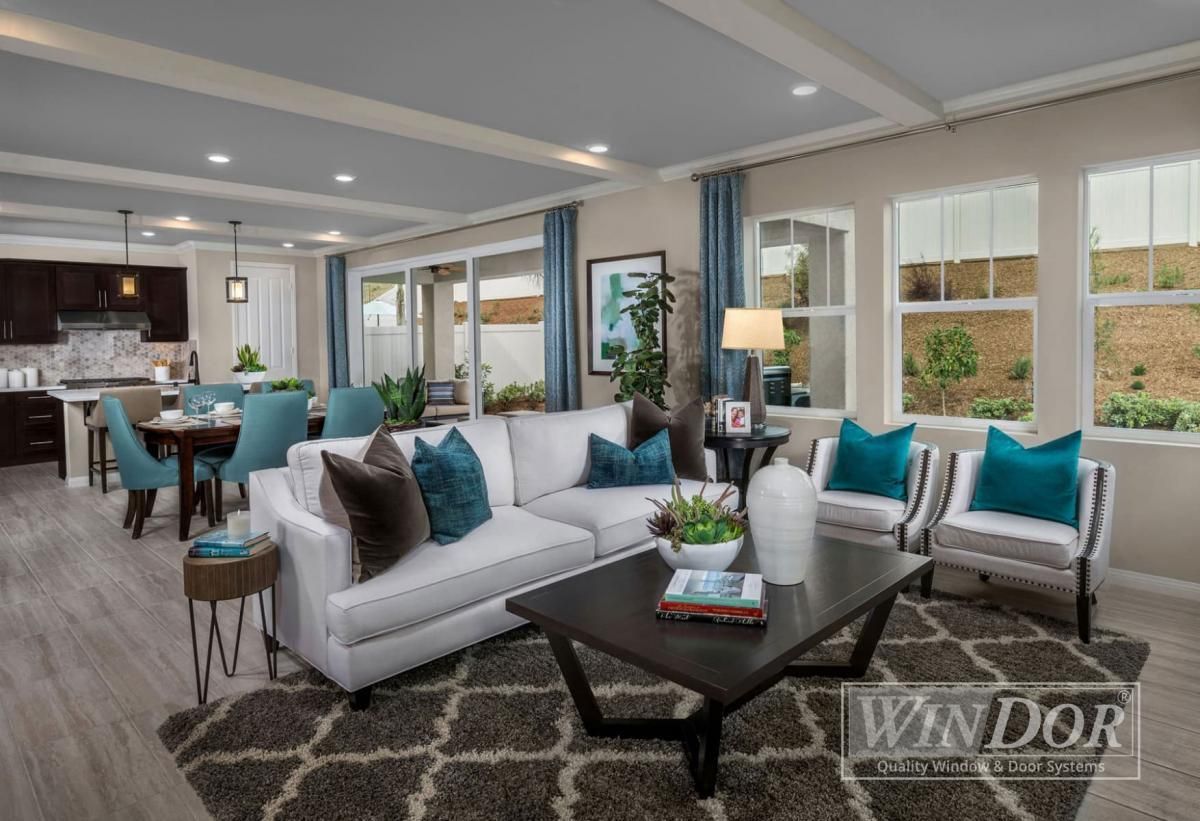 Modern open-concept living room with white sofas, teal pillows, and large windows overlooking a patio.