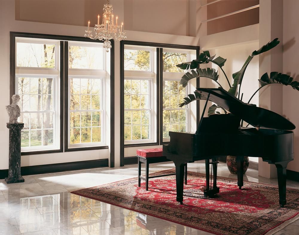 Elegant living room with a grand piano, red rug, chandelier, and tall windows with sunlight.