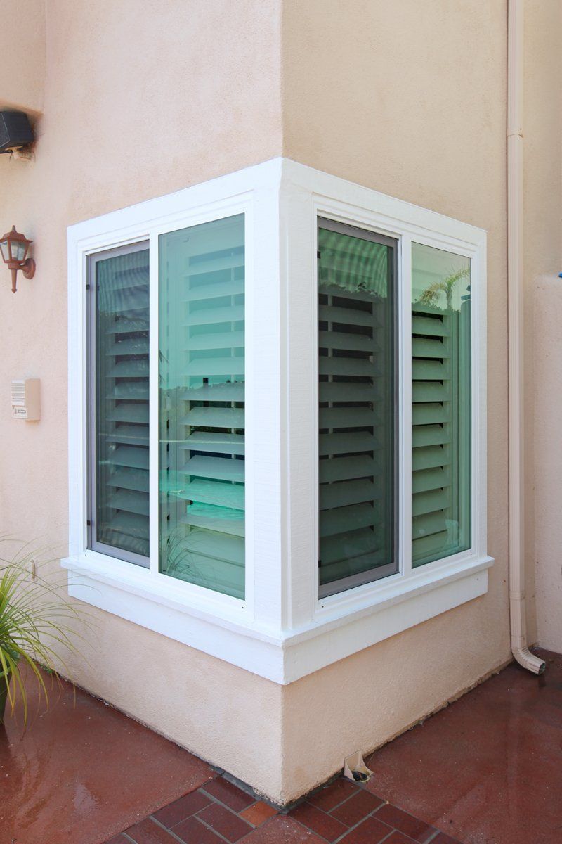 White bay window with green shutters on a beige stucco house corner, above a red tiled patio
