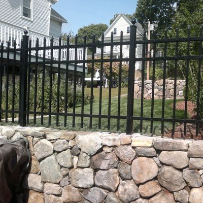 Black metal fence atop a stone wall, surrounding a grassy yard, near a house.