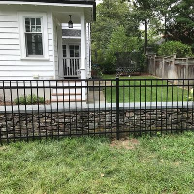 Black metal fence atop a stone wall bordering a grassy yard and white house.