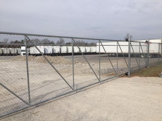 Chain-link fence in front of a gravel lot with parked semi-truck trailers, under a cloudy sky.