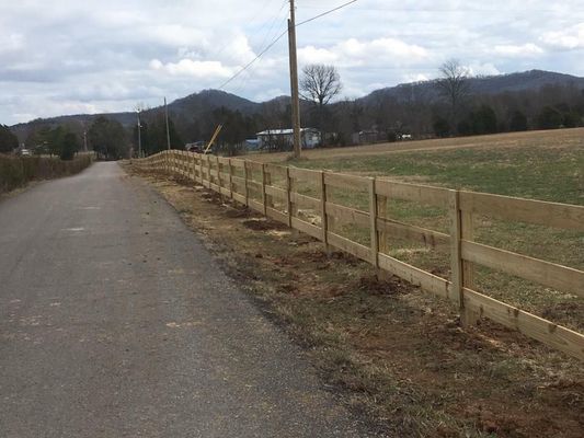A wooden fence lines a rural road, beside a field with mountains in the background.