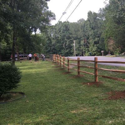 Wooden split-rail fence lining a green lawn with people working; trees and road in the background.