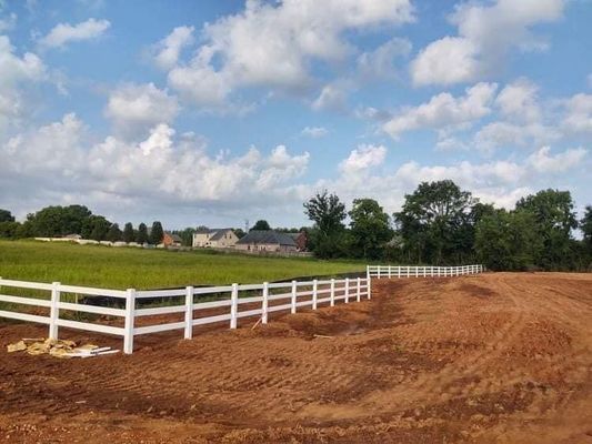 White fence borders a field of green grass and a dirt area, with a house in the background under a cloudy sky.