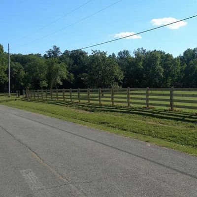 Roadside view of a wooden fence bordering a green field, trees in the background, and a clear blue sky.