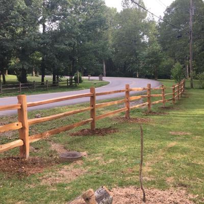 Wooden split-rail fence alongside a curving road, with grass and trees in the background.