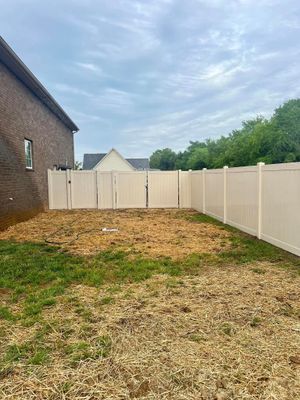 A tan vinyl fence encloses a bare yard next to a brick house on an overcast day.