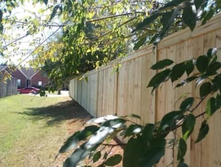 Wooden fence in a grassy yard, partially obscured by tree branches, with a house in the background.