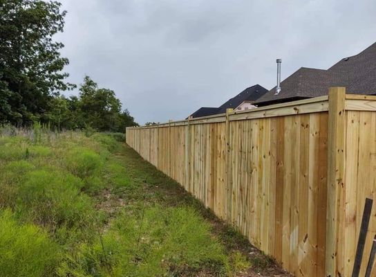 Wooden fence along overgrown grassy area, separating homes. Cloudy sky.