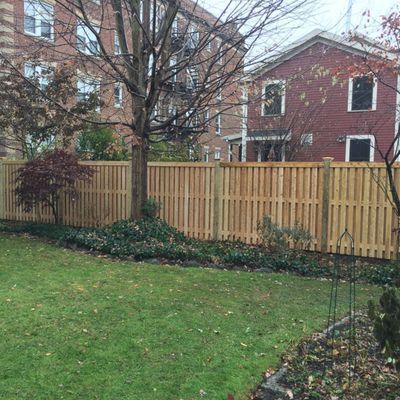 Wooden fence around a backyard with green grass, small tree, and two brick buildings.