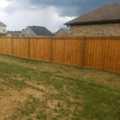 Wooden fence in a grassy backyard with houses in the background under a cloudy sky.