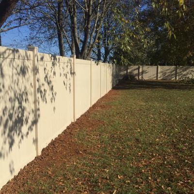 Beige vinyl fence bordering a grassy yard with a dirt patch and trees in the background.