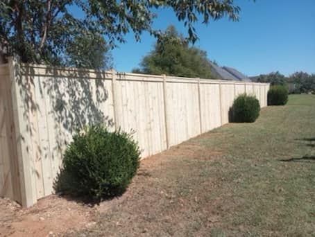 Wooden fence alongside a grassy yard with green bushes and a clear blue sky.