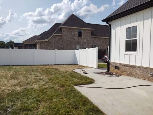 A backyard with a white fence, concrete patio, and a house. The grass is mostly brown and the sky is partly cloudy.