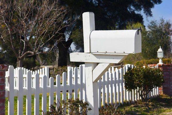 White mailbox on a white picket fence in front of a brick wall and trees.