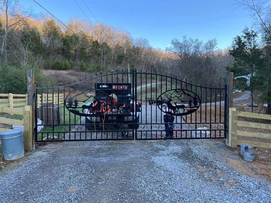 Black iron gates with decorative cutouts open to a gravel driveway. Wooden fences flank the gates.