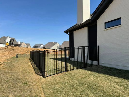 Black metal fence around a grassy area next to a white brick house with a black shutter and a chimney.
