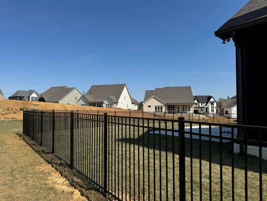 Black metal fence in a grassy yard, with houses and a blue sky in the background.