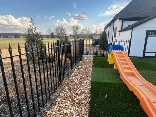 Black iron fence along a stone path next to a house with a playground slide on turf.