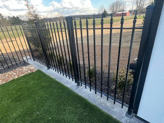 Black metal fence bordering green artificial grass, with a cement base.
