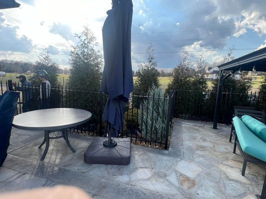 Patio with table, closed umbrella, green bushes, black fence, and seating under a cloudy sky.