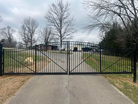 Black metal gates closed on a driveway, leading to a building with bare trees in the background.