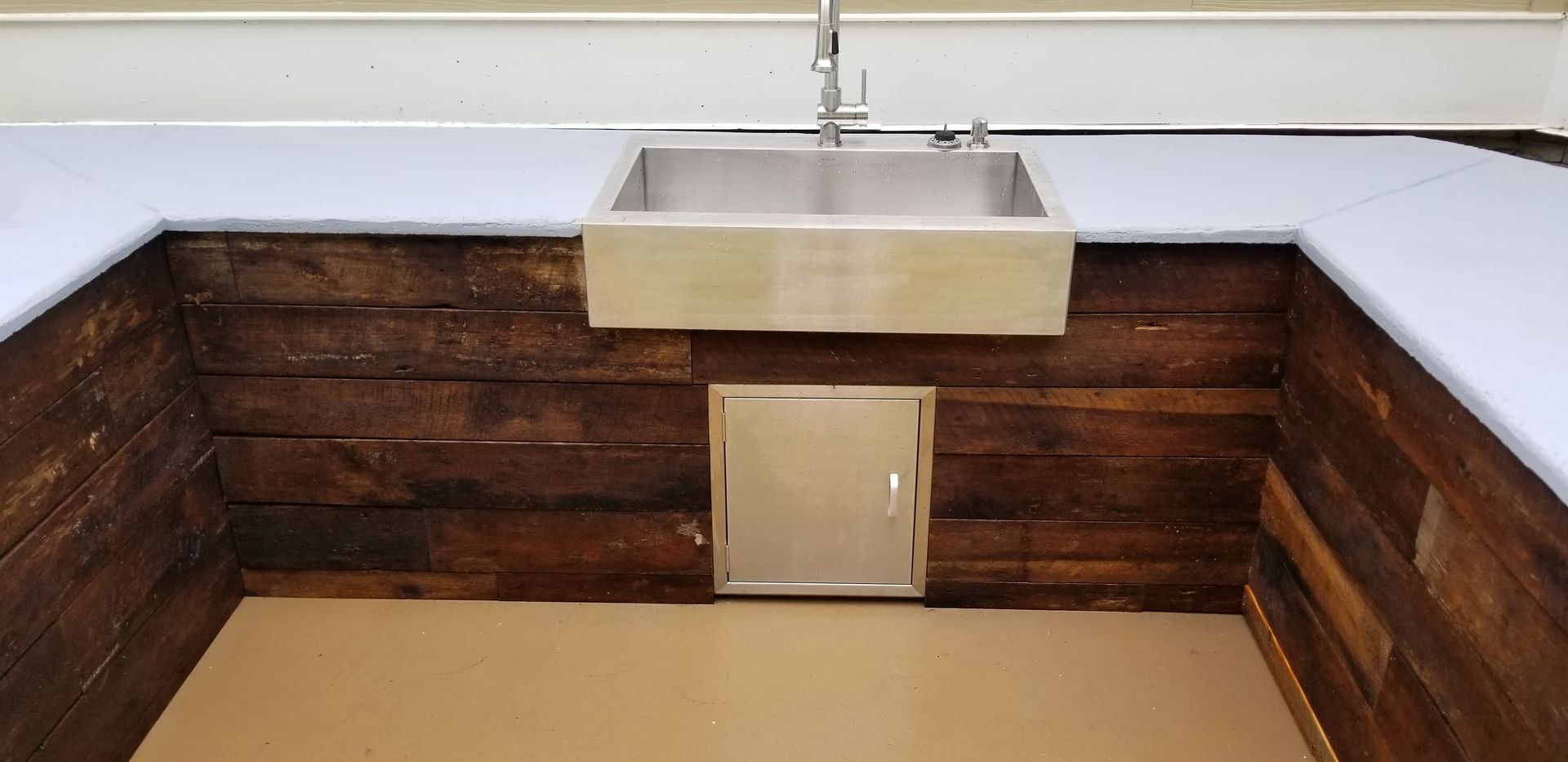 An outdoor kitchen with a stainless steel sink, wooden paneling, and a small stainless steel door. A light-colored countertop extends around the sink.