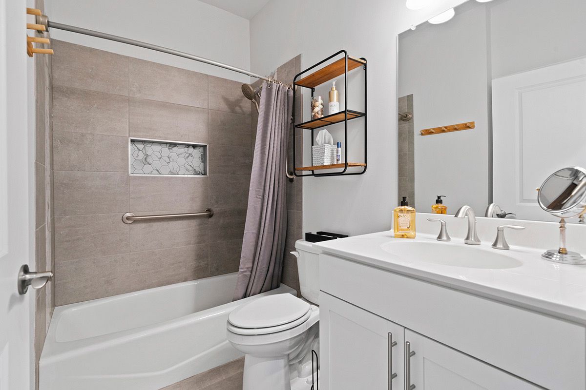 A small, modern bathroom with a bathtub, toilet, and vanity.  Gray tile, white fixtures, and a metal shelf give a minimalist feel.