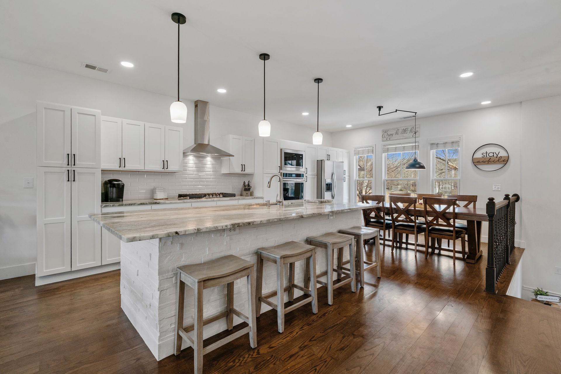 Modern kitchen with a large island, seating, and a dining area with a wooden table and chairs. White cabinets and stainless steel appliances.