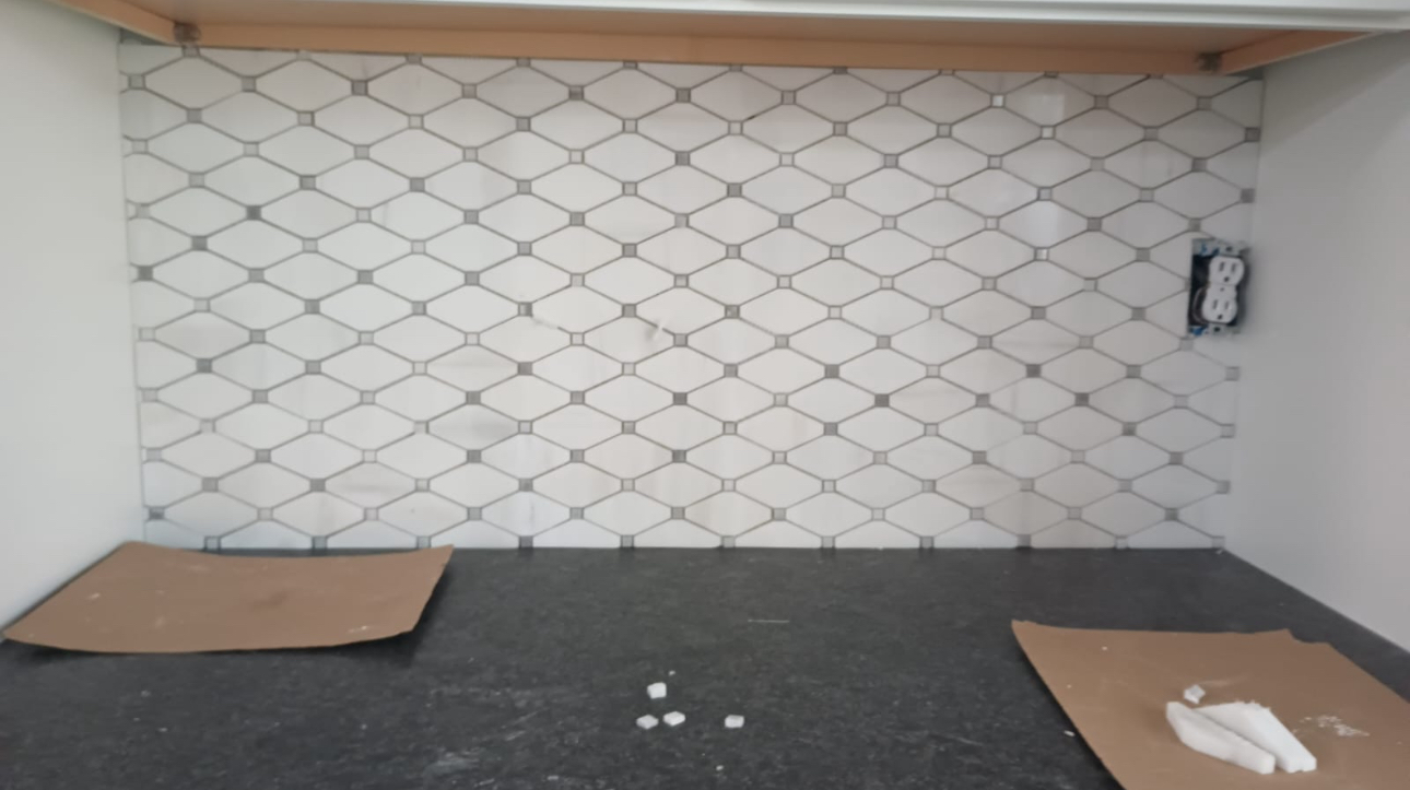 Kitchen backsplash tiling in progress. White and gray patterned tiles are installed above a dark countertop, with cardboard and supplies below.