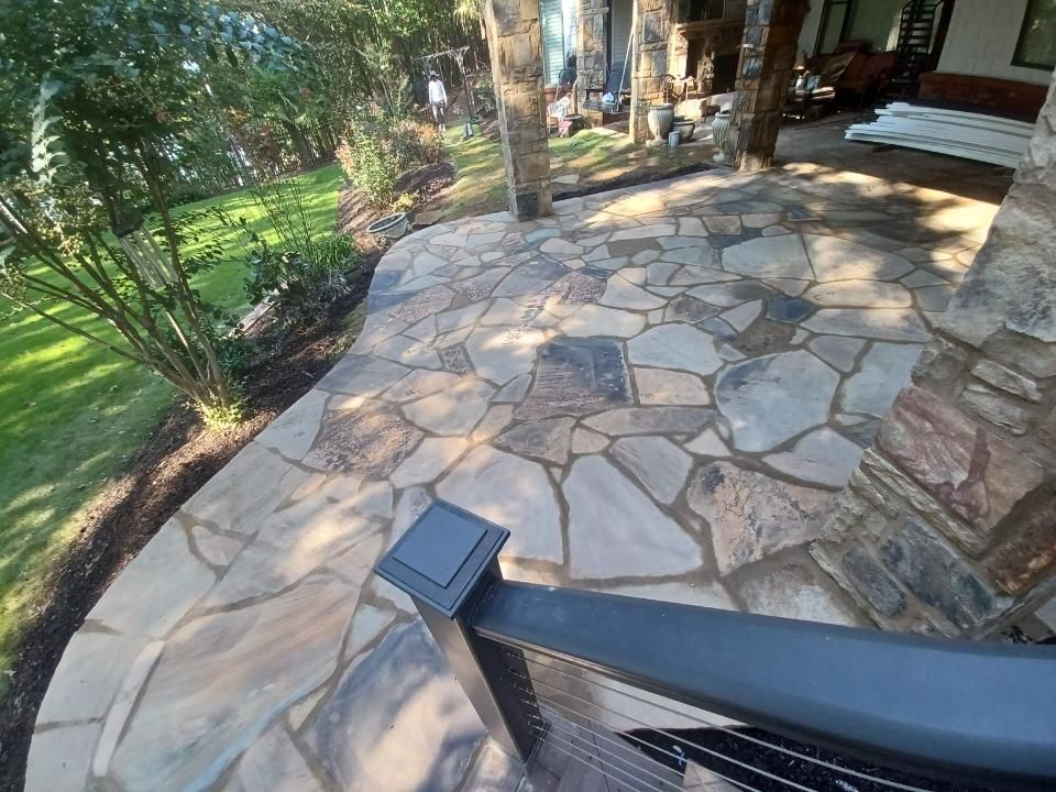 Stone patio with a black railing in the foreground, leading toward a covered porch area with stone columns and lush green grass.