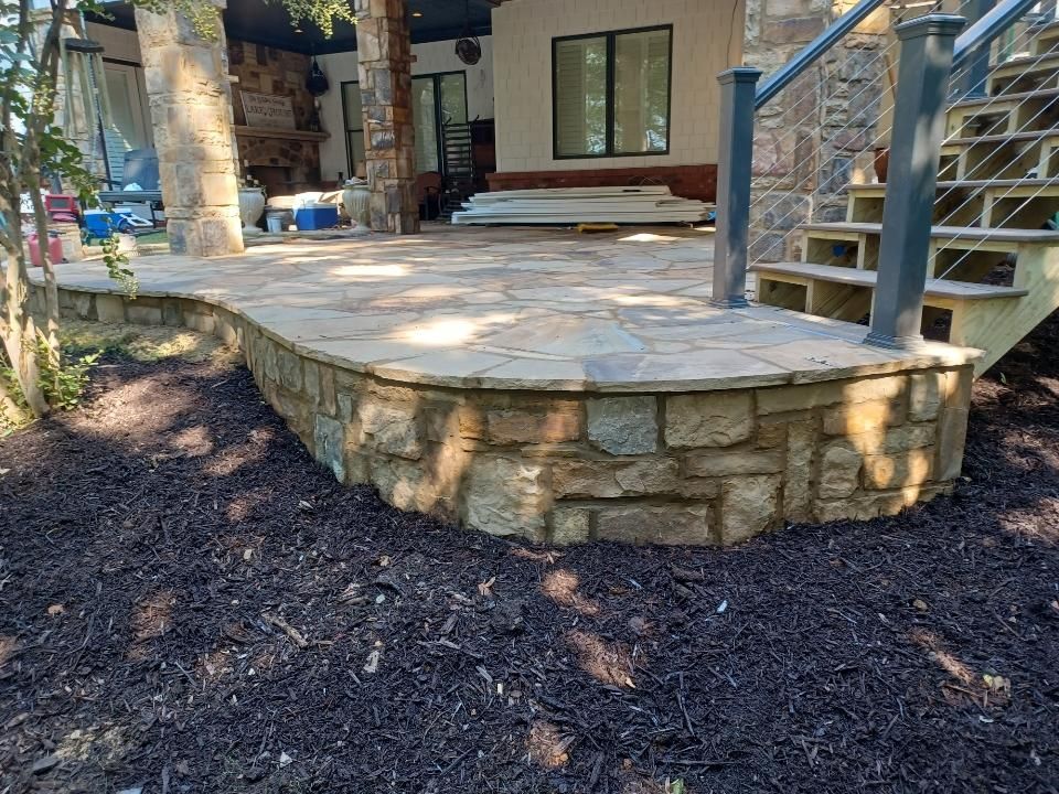 A stone patio with a curved retaining wall sits in front of a house with steps and a covered area, surrounded by mulch and trees.