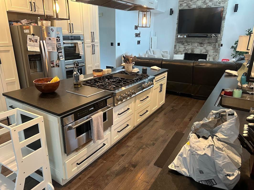 Spacious kitchen with large island containing a stovetop, oven, and cabinets. Wooden floors, white cabinets, and a stainless steel refrigerator.