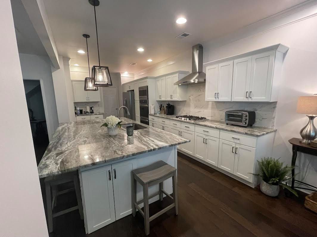 A bright kitchen with white cabinets, a large granite island, and stainless steel appliances. A stool sits at the island.