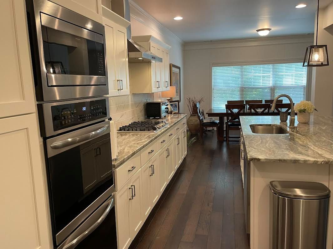Narrow kitchen with white cabinets, stainless steel appliances, and granite countertops. Dark wood floors lead to a dining area with a table and chairs.