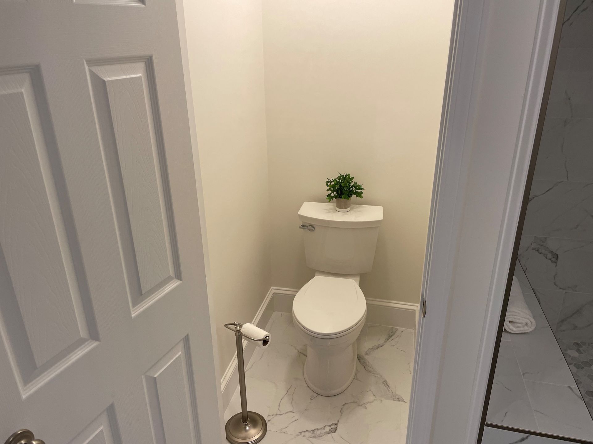 A small white bathroom with a toilet and a plant on the tank. The floor is white marble-patterned tile.