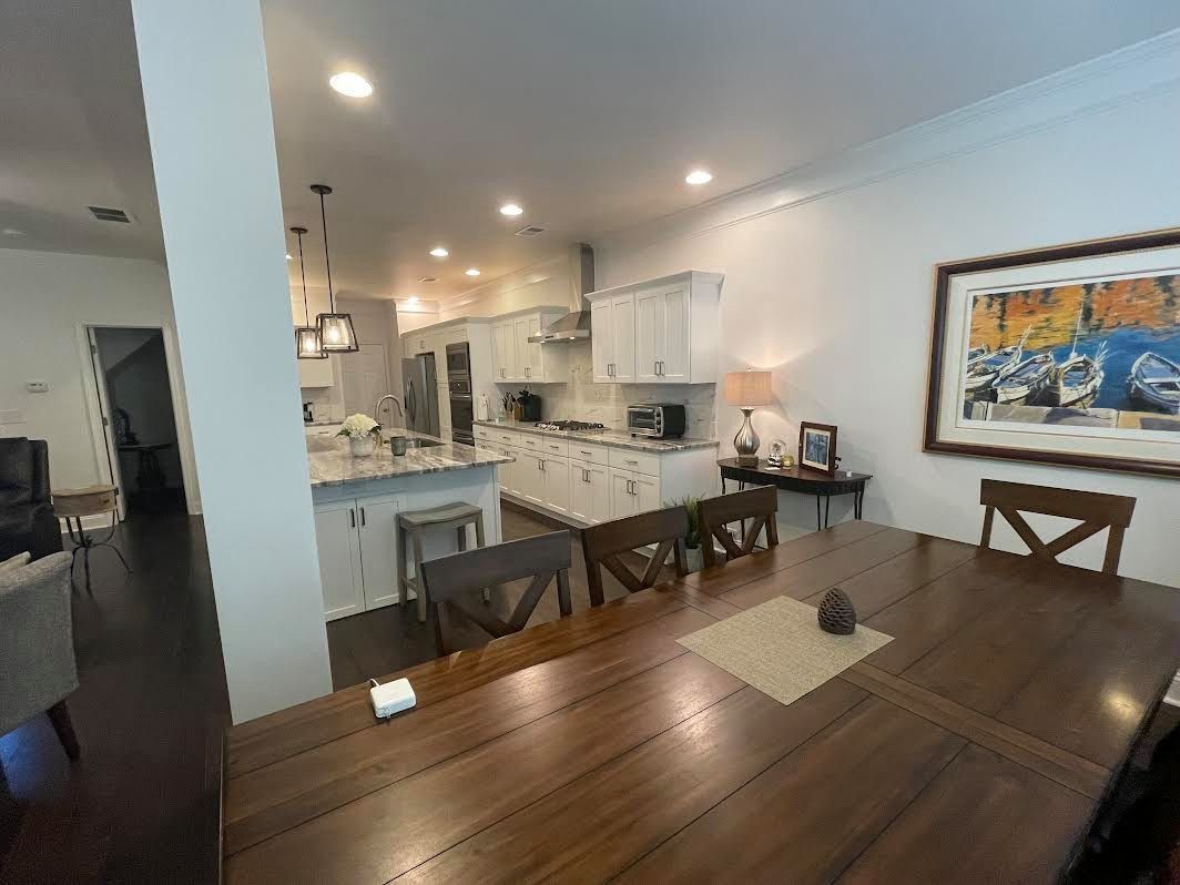 A bright kitchen and dining area with a long wooden table, white cabinets, and a painting on the wall.