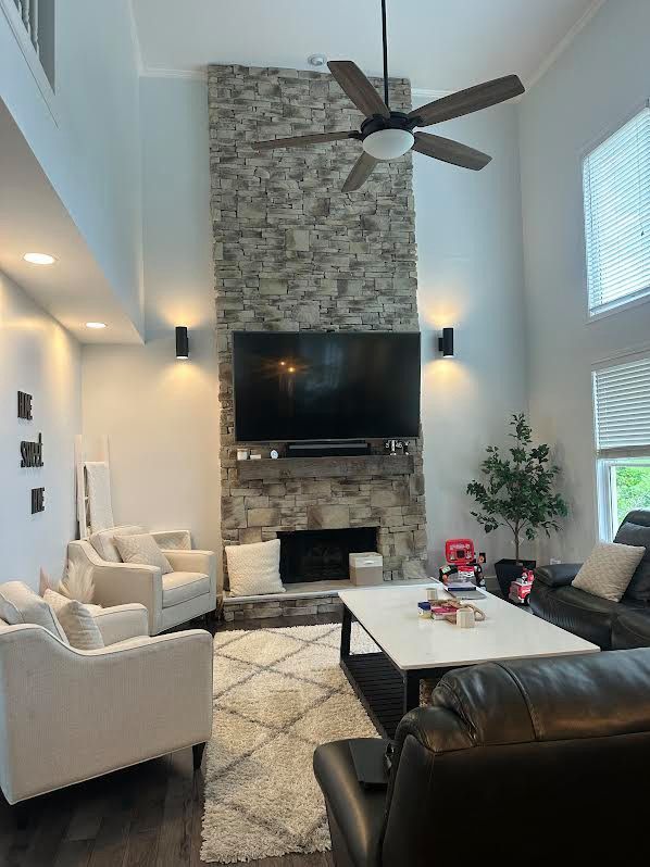 Living room with a stone fireplace, large TV, two black sconces, and two white armchairs. A ceiling fan hangs overhead, and a black leather couch sits nearby.