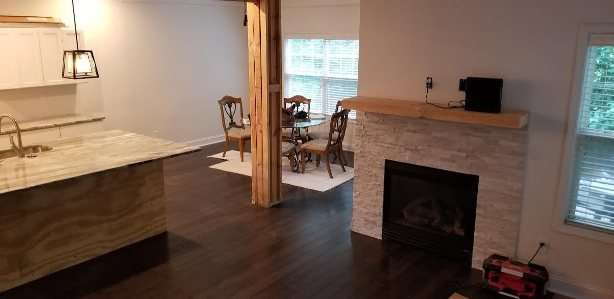 A living room with a kitchen. It has dark hardwood floors, a fireplace with white stone, a wooden pillar, and antique furniture.