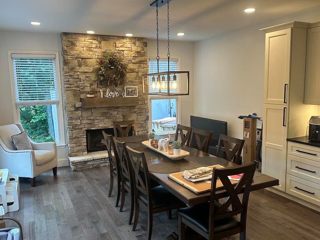 A dining room with a large wooden table, stone fireplace, and chairs. Window with blinds on the left, light fixtures above the table.