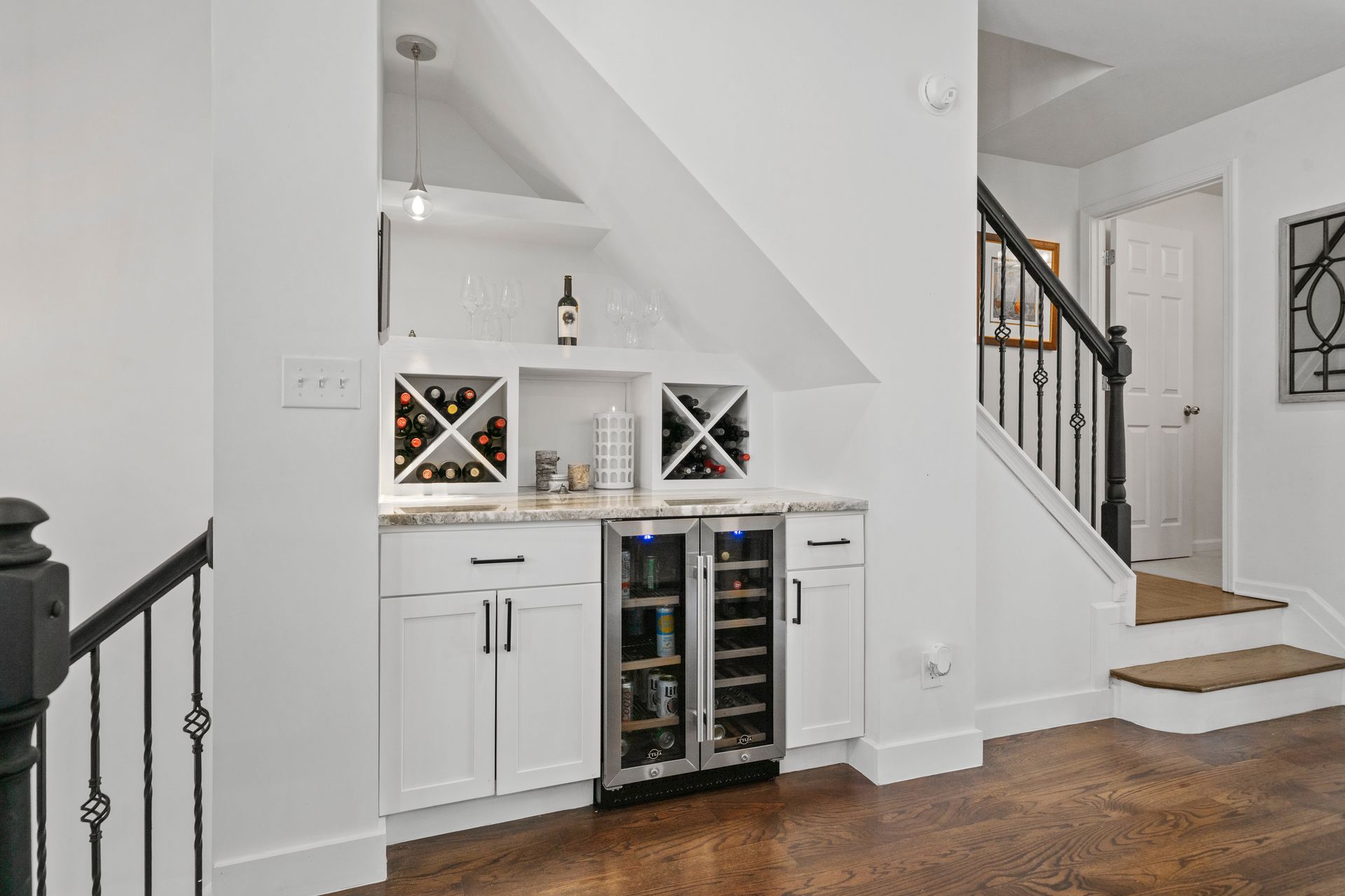 Wine bar built into the wall under a staircase. White cabinets, granite countertop, wine fridge, and racks.