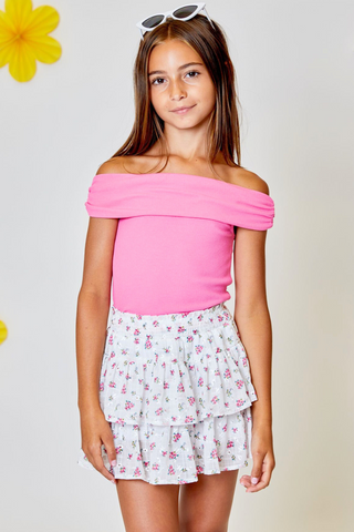 A young girl wearing a pink off the shoulder top and a white floral skirt.