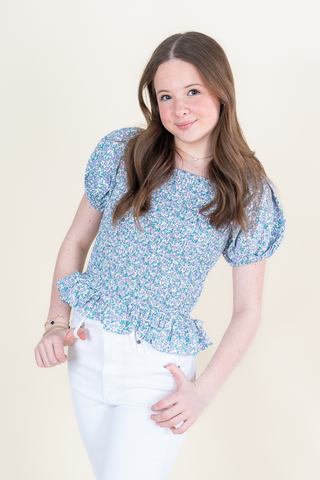 A young girl is wearing a blue floral top and white pants.
