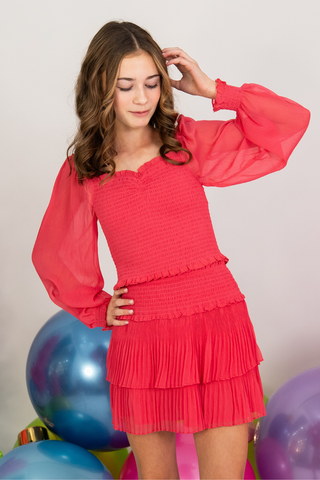 Person in a bright pink dress with sheer sleeves and a tiered skirt, standing near colorful balloons.
