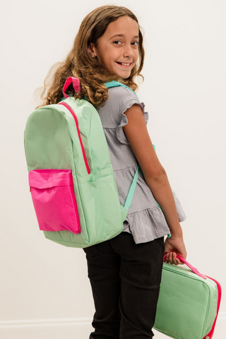 A young girl is carrying a green backpack and a pink lunch bag.